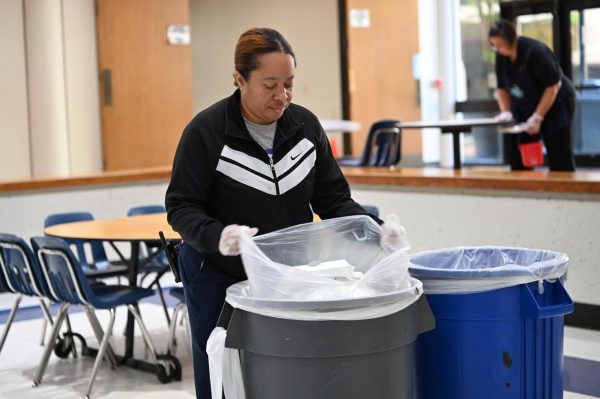 Custodian Latonia Wynn takes out a load of trash following the lunch shift with custodian Arin Davies in the background, wiping off tables to prepare for the next lunch.